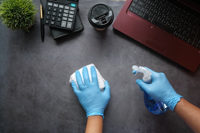Hands in blue gloves using a spray bottle and towel to wipe down a desk